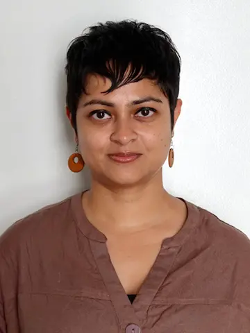 A woman with short dark hair wearing a brown top and brown earrings against a pale background.